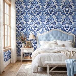 May include: A bedroom featuring wallpaper with a blue and white damask pattern. A light blue tufted headboard sits behind a bed with white bedding and a light blue throw. A white nightstand holds a lamp, a framed picture, and books.