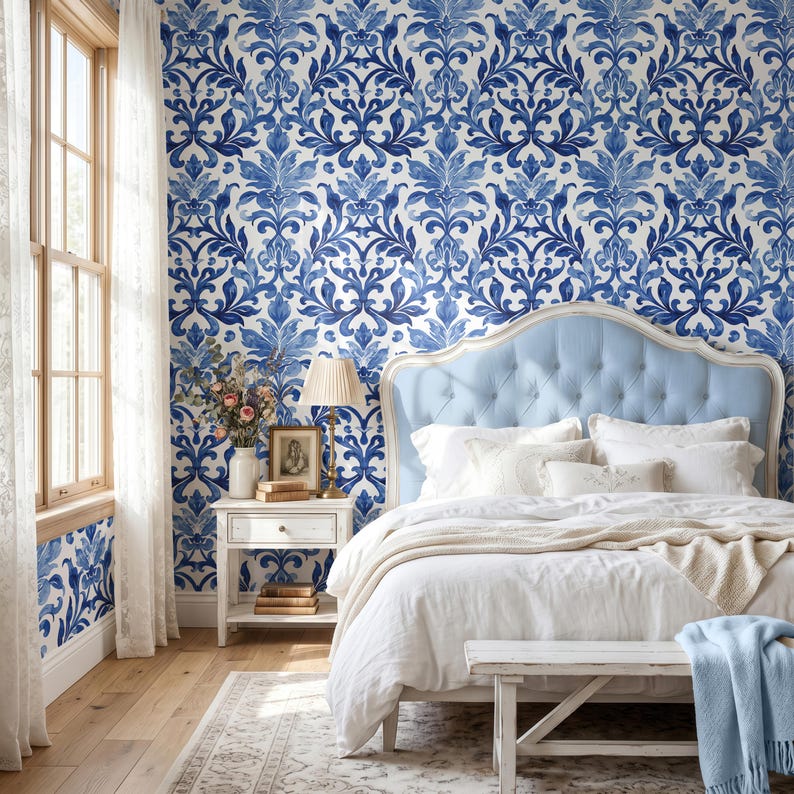 May include: A bedroom featuring wallpaper with a blue and white damask pattern. A light blue tufted headboard sits behind a bed with white bedding and a light blue throw. A white nightstand holds a lamp, a framed picture, and books.