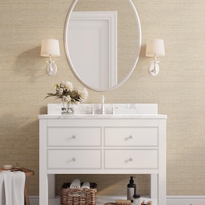 May include: A white bathroom vanity with four drawers, a marble countertop, and an oval mirror. The vanity has a lower shelf with a wicker basket, toiletries, and a brush. Two wall sconces flank the mirror. A rug and a small wooden stool are in the foreground.