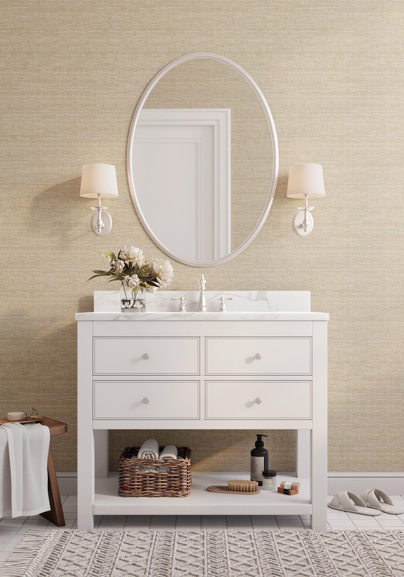 May include: A white bathroom vanity with four drawers, a marble countertop, and an oval mirror. The vanity has a lower shelf with a wicker basket, toiletries, and a brush. Two wall sconces flank the mirror. A rug and a small wooden stool are in the foreground.