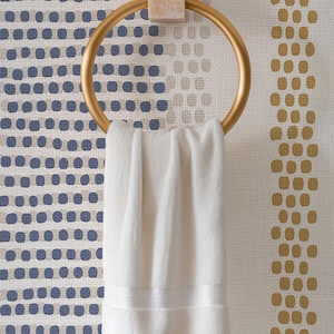 Op de afbeelding: Een goudkleurige handdoekring met een witte handdoek. De ring is gemonteerd op een lichtgekleurd, getextureerd blok. De achtergrond heeft een behang met blauwe en gouden stippen.