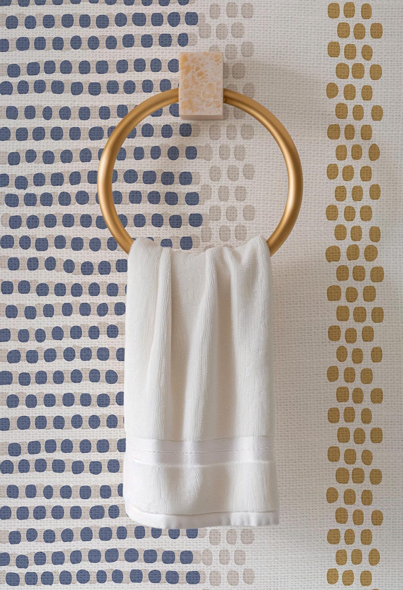 Op de afbeelding: Een goudkleurige handdoekring met een witte handdoek. De ring is gemonteerd op een lichtgekleurd, getextureerd blok. De achtergrond heeft een behang met blauwe en gouden stippen.