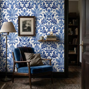 May include: A blue and white damask patterned wallpaper covers the wall behind a dark wood armchair with blue velvet upholstery. A floor lamp with a pleated shade, a framed black and white print, and a small wooden shelf with books are visible.