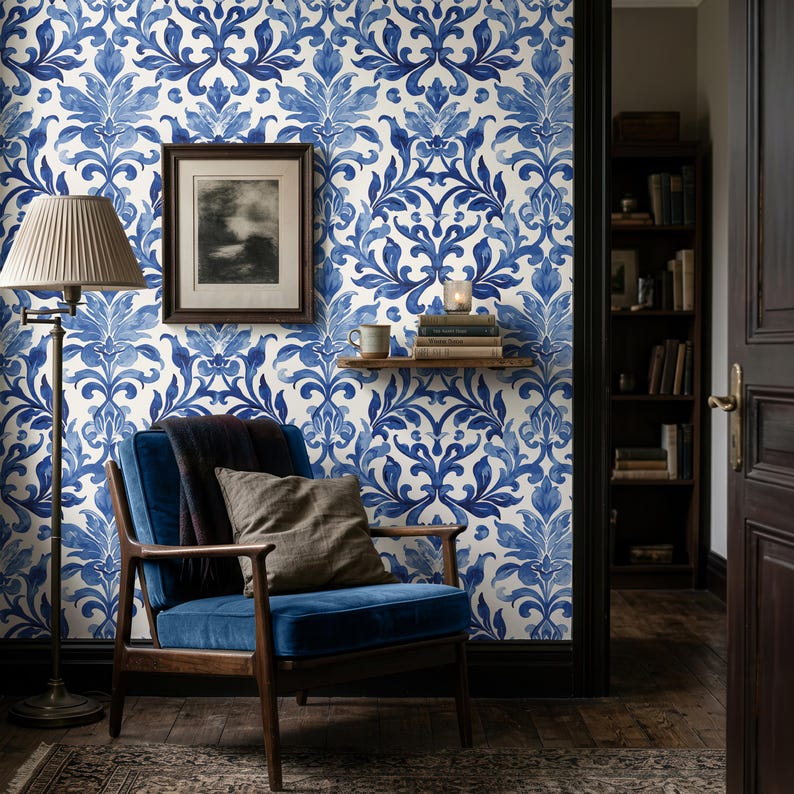 May include: A blue and white damask patterned wallpaper covers the wall behind a dark wood armchair with blue velvet upholstery. A floor lamp with a pleated shade, a framed black and white print, and a small wooden shelf with books are visible.