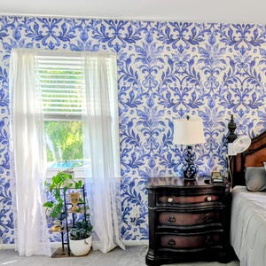 May include: A room interior featuring wallpaper with a blue and white damask pattern. A window is covered by sheer white curtains. A dark wood three-drawer nightstand holds a lamp with a white shade. A metal plant stand holds a potted green plant.