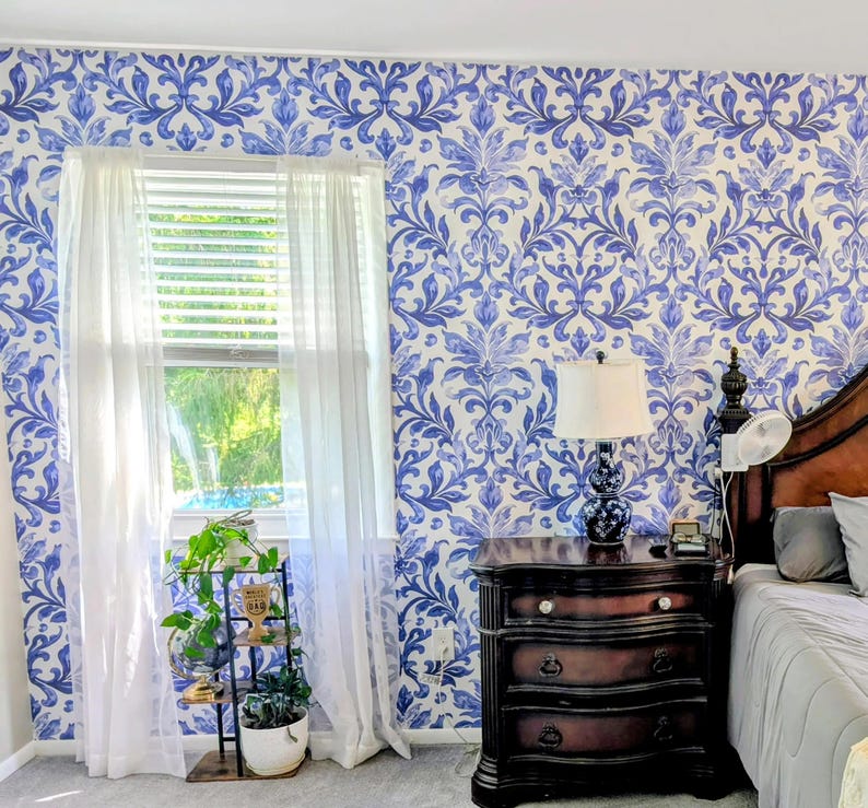 May include: A room interior featuring wallpaper with a blue and white damask pattern. A window is covered by sheer white curtains. A dark wood three-drawer nightstand holds a lamp with a white shade. A metal plant stand holds a potted green plant.