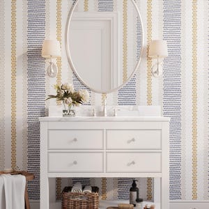 Op de afbeelding: Witte badkamermeubel met een marmeren blad, ovale spiegel en wandkandelaars. Het behang heeft verticale strepen van blauwe, gouden en witte stippen. Een gevlochten mand, borstel en zeep staan op de plank.