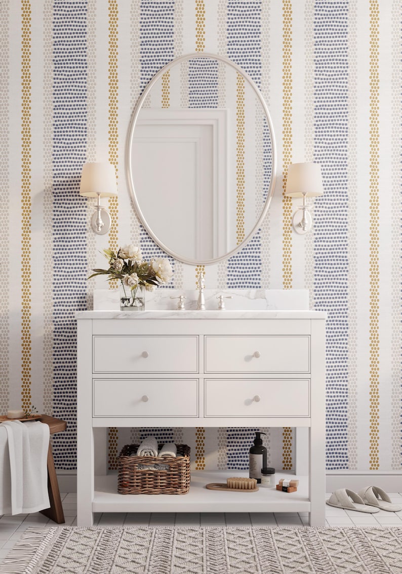Op de afbeelding: Witte badkamermeubel met een marmeren blad, ovale spiegel en wandkandelaars. Het behang heeft verticale strepen van blauwe, gouden en witte stippen. Een gevlochten mand, borstel en zeep staan op de plank.
