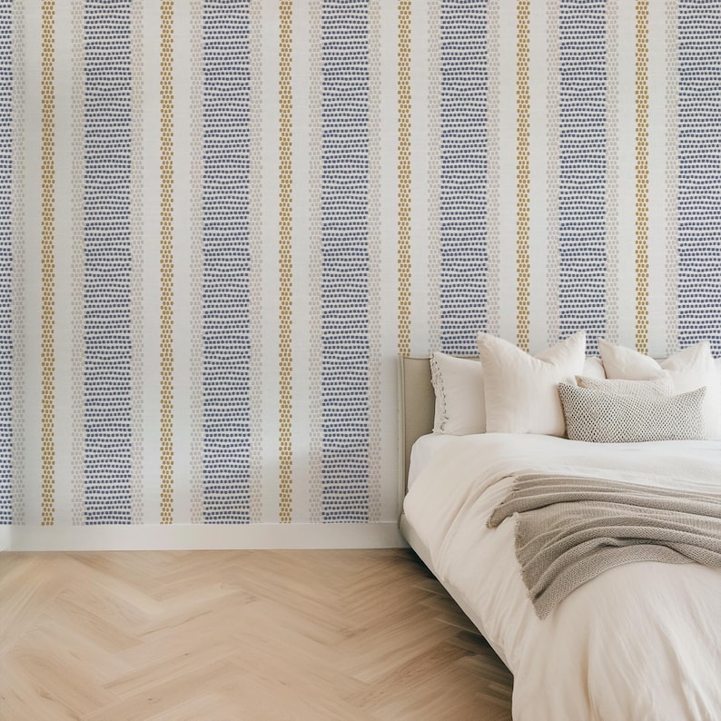 Op de afbeelding: Een slaapkamer met behang met een patroon en een bed. Het behang heeft verticale strepen van witte, blauwe en gouden stippen. Het bed heeft wit beddengoed, kussens en een grijze deken. De vloer is lichtgekleurd hout.