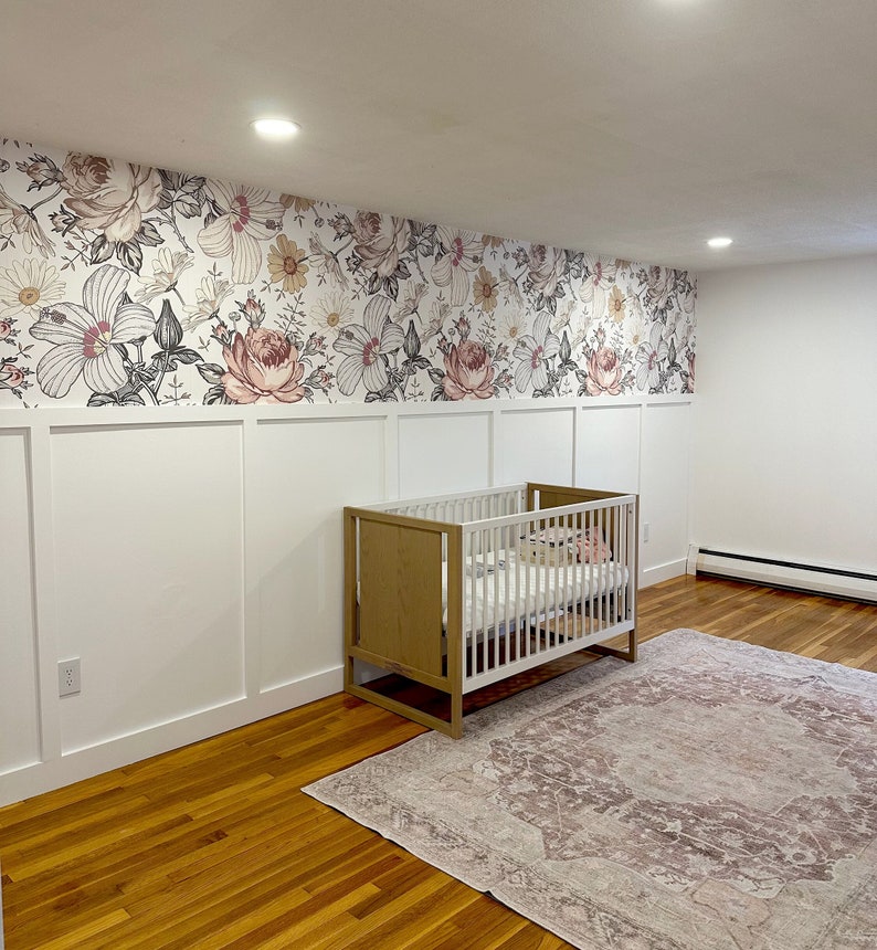May include: A white crib with a wooden frame and a floral patterned wallpaper behind it. A pink and white rug is on the floor in front of the crib.