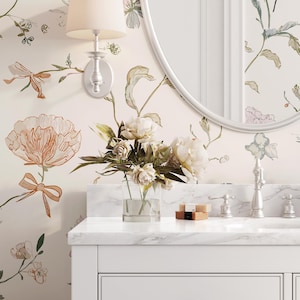 Puede incluir: Un mueble de baño blanco con encimera de mármol, grifo plateado y espejo redondo. La pared presenta papel pintado floral con detalles melocotón y verde. Un jarrón de cristal contiene flores blancas.
