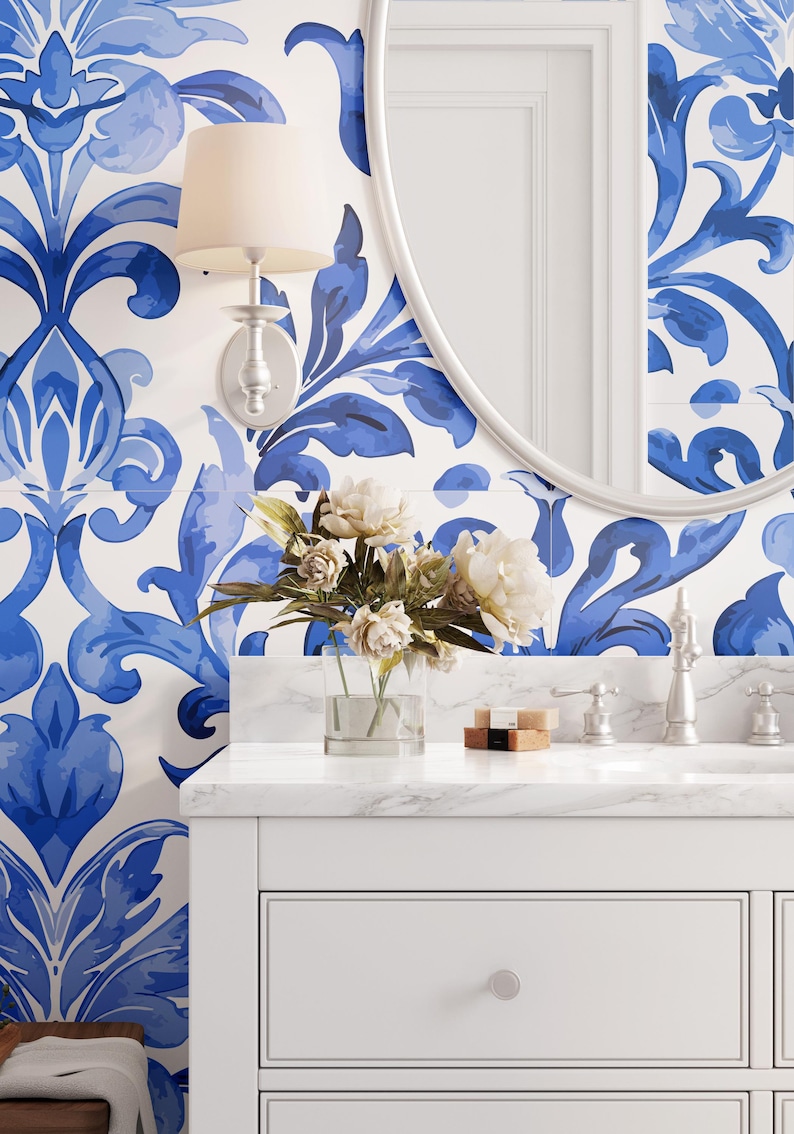 May include: A bathroom vanity with a white marble countertop, a white cabinet, and a round mirror. The wall features blue and white damask patterned wallpaper. A glass vase with white flowers, a wall sconce, and soap bars are on the counter.