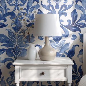 May include: A white nightstand with a single drawer and a small round knob, topped with a beige table lamp featuring a white drum shade and a small white vase containing thin dried branches, set against a blue and white damask patterned wallpaper.
