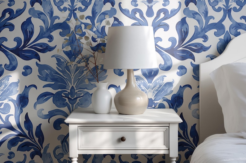May include: A white nightstand with a single drawer and a small round knob, topped with a beige table lamp featuring a white drum shade and a small white vase containing thin dried branches, set against a blue and white damask patterned wallpaper.