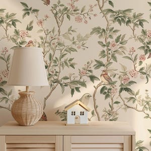 May include: A beige wallpaper with a floral pattern featuring green leaves, pink flowers, and small brown birds. A wooden cabinet with two doors is in the foreground. A small white house and a wicker lamp are on top of the cabinet.