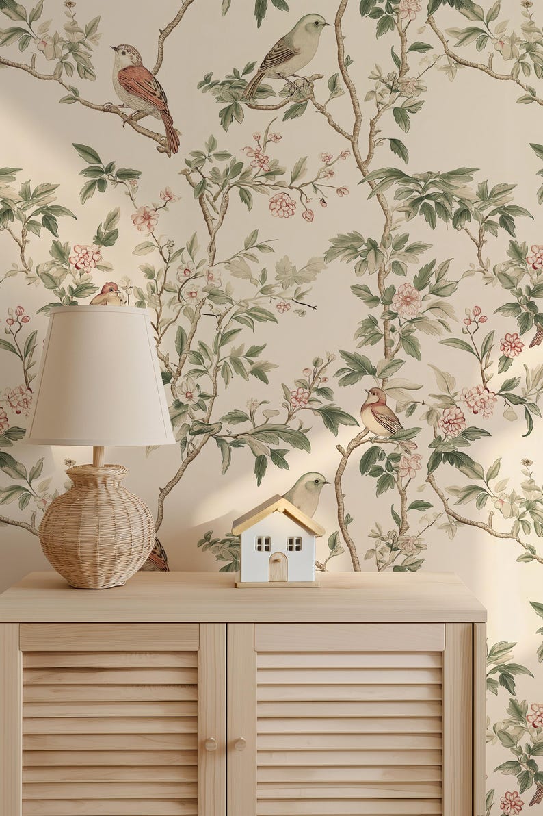 May include: A beige wallpaper with a floral pattern featuring green leaves, pink flowers, and small brown birds. A wooden cabinet with two doors is in the foreground. A small white house and a wicker lamp are on top of the cabinet.
