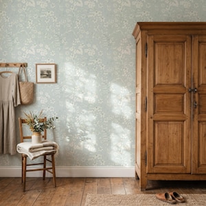 May include: A tall wooden wardrobe with double doors stands next to a wooden chair holding a folded blanket and a vase of dried flowers. A linen dress hangs on a wall hook beside a framed picture, all set against light blue floral wallpaper.