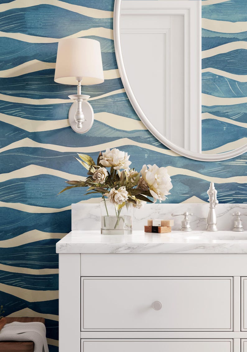 May include: A white bathroom vanity with a marble countertop. The vanity has two drawers and a single knob on the top drawer. A clear glass vase with white flowers sits on the countertop. A silver wall sconce with a white shade is mounted on the wall behind the vanity. A round, white mirror is mounted on the wall above the vanity. The wall behind the vanity is covered in blue and cream wallpaper with a wavy pattern.