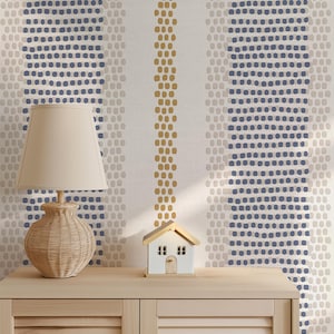 Op de afbeelding: Een beige rotan lamp met een cr&egrave;mekleurige lampenkap staat op een licht houten kast. Een klein wit huismodel met een bruin dak staat naast de lamp. De achtergrond is een behang met verticale strepen van blauwe, beige en gouden stippen.