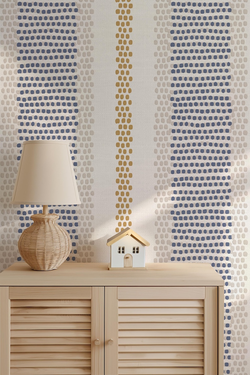 Op de afbeelding: Een beige rotan lamp met een cr&egrave;mekleurige lampenkap staat op een licht houten kast. Een klein wit huismodel met een bruin dak staat naast de lamp. De achtergrond is een behang met verticale strepen van blauwe, beige en gouden stippen.