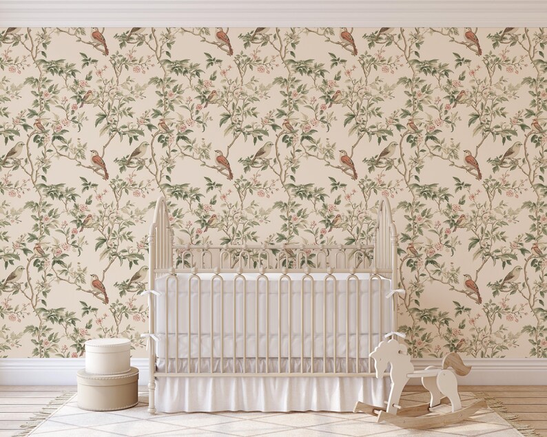 May include: A white cot with a white frilled skirt and a white rocking horse in front of it. The wall behind the cot is covered in a beige wallpaper with a repeating pattern of brown birds and green leaves.