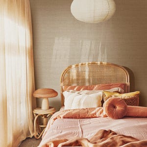 May include: A bedroom scene featuring a bed with a woven headboard, adorned with pink and peach bedding and pillows. A small mushroom-shaped side table and a paper lantern hang from the ceiling. The room has a warm, inviting atmosphere.