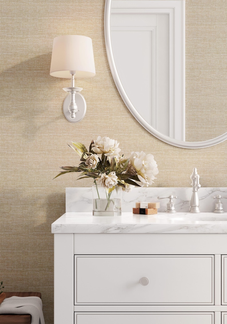 May include: A bathroom scene featuring a white vanity with a marble countertop, a clear vase of white flowers, and a silver faucet. A wall-mounted light fixture with a beige lampshade is above the vanity, and a round mirror is on the wall.