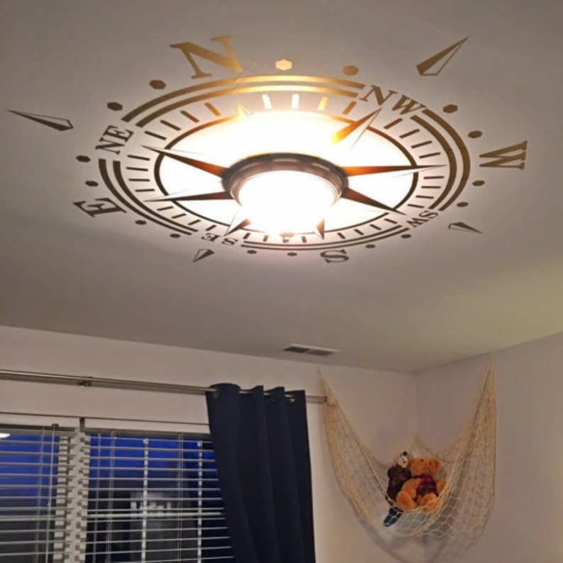 May include: A gold compass design on a white ceiling with a light fixture in the center. The compass points are labeled N, NE, E, SE, S, SW, W, and NW.