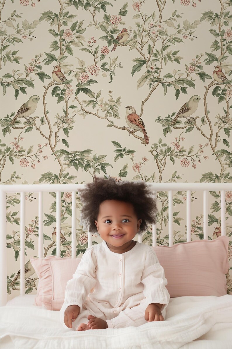 May include: A young child sits on a white cot with a floral wallpaper featuring a repeating pattern of green leaves, pink flowers, and brown birds. The child is wearing a white long-sleeved onesie.