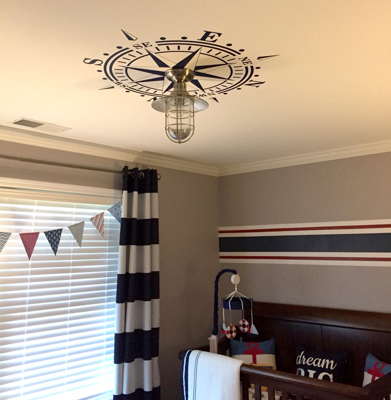 May include: A nursery room with a white ceiling featuring a blue and white compass decal. The room has a white and blue striped curtain, a white crib with a navy blue and red striped bumper, and a white and blue striped blanket. The wall is painted a light gray with a navy blue and red stripe.