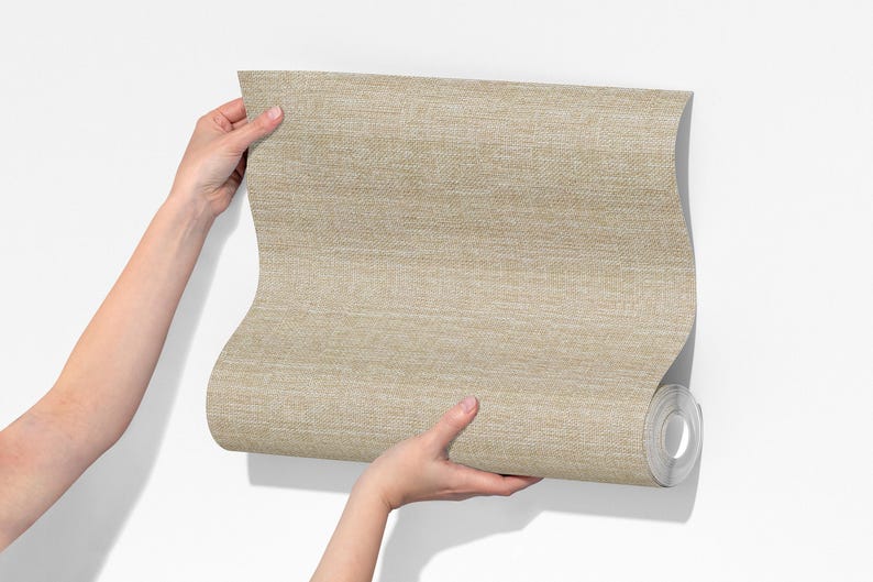 May include: A roll of beige wallpaper with a woven texture is held against a white wall. The wallpaper is a neutral color, and the texture is visible. The roll is partially unrolled, revealing the paper's interior.