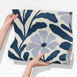 May include: A roll of wallpaper featuring a repeating floral pattern. The design includes large, stylized flowers and leaves in shades of blue and gray against a cream background. The wallpaper is held against a white wall.