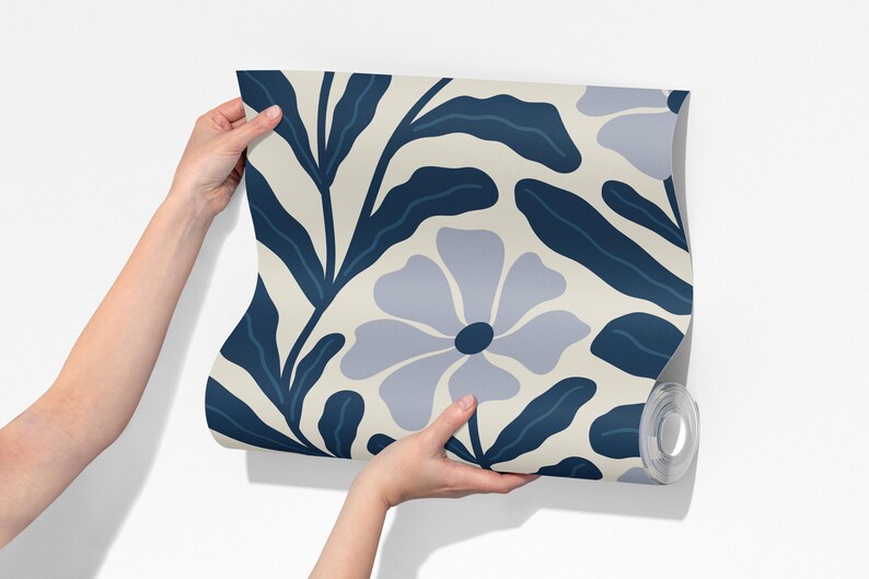 May include: A roll of wallpaper featuring a repeating floral pattern. The design includes large, stylized flowers and leaves in shades of blue and gray against a cream background. The wallpaper is held against a white wall.
