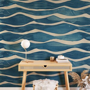 May include: A room with a blue and beige wave pattern wallpaper. A light wood desk with a small drawer, a white lamp, and a fluffy stool are in front of the wall. A vase with dried plants is on the right.