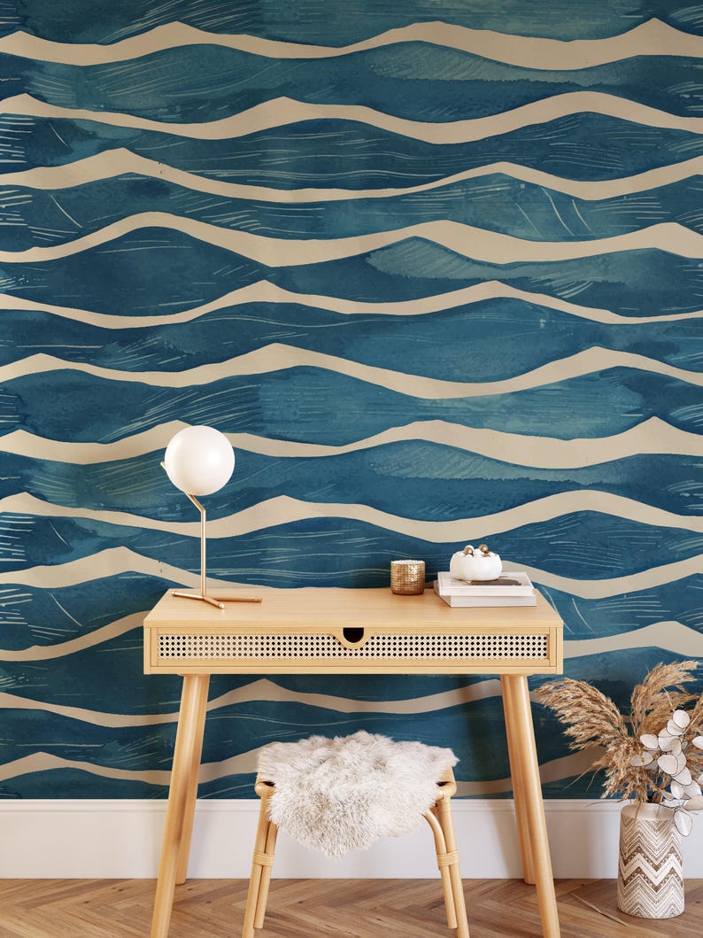 May include: A room with a blue and beige wave pattern wallpaper. A light wood desk with a small drawer, a white lamp, and a fluffy stool are in front of the wall. A vase with dried plants is on the right.