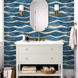 May include: A bathroom with a white vanity, gold fixtures, and a blue and white wave-patterned wallpaper. The vanity has two drawers and a shelf with baskets of towels. A gold-framed mirror and sconces complete the design.