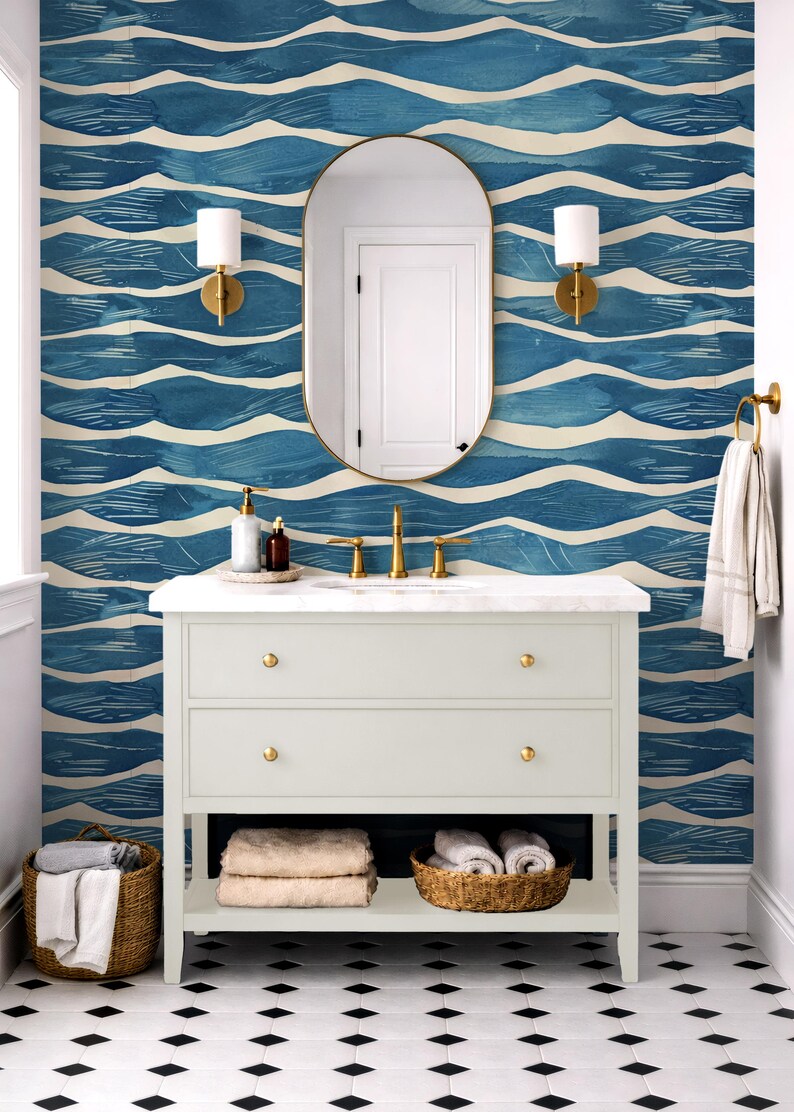 May include: A bathroom with a white vanity, gold fixtures, and a blue and white wave-patterned wallpaper. The vanity has two drawers and a shelf with baskets of towels. A gold-framed mirror and sconces complete the design.