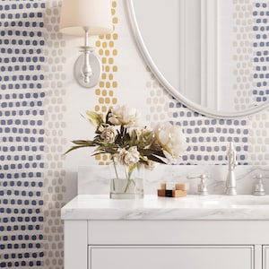 Op de afbeelding: Badkamerinterieur met een witte wastafel, marmeren aanrechtblad en zilveren kranen. Het behang heeft een stippenpatroon in blauw, goud en beige. Een heldere vaas bevat witte bloemen.