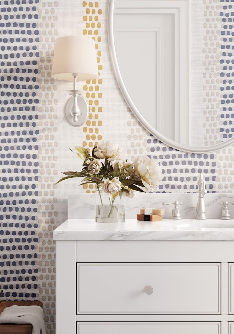 Op de afbeelding: Badkamerinterieur met een witte wastafel, marmeren aanrechtblad en zilveren kranen. Het behang heeft een stippenpatroon in blauw, goud en beige. Een heldere vaas bevat witte bloemen.