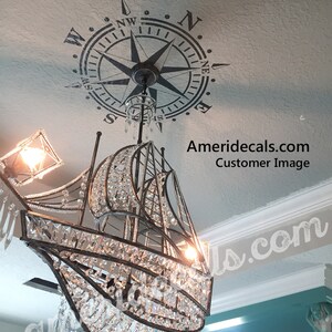 May include: A crystal chandelier shaped like a sailboat hangs from a ceiling with a compass stencil. The compass points north and has the letters N, W, E, and S around the edge.
