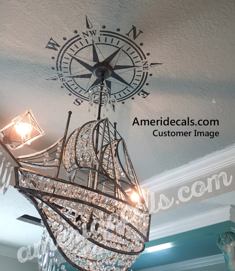 May include: A crystal chandelier shaped like a sailboat hangs from a ceiling with a compass stencil. The compass points north and has the letters N, W, E, and S around the edge.