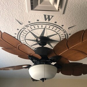 May include: A ceiling fan with brown wooden blades in the shape of palm leaves. The fan is installed on a white ceiling with a compass stencil design. The compass design features a black and gray compass rose with the letters N, W, S, and E.