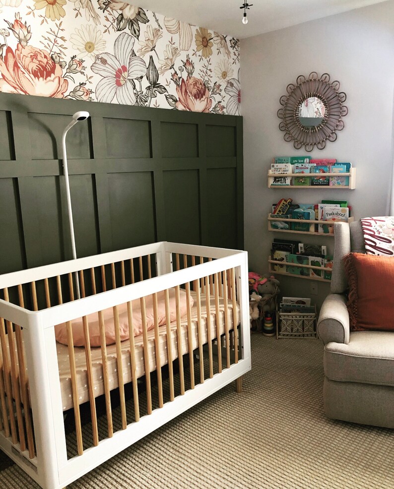 May include: A white crib with wooden slats and a pink and white patterned sheet. The crib is in a nursery with green wainscoting and floral wallpaper. There is a gray armchair with a red throw pillow and a bookshelf with children's books.