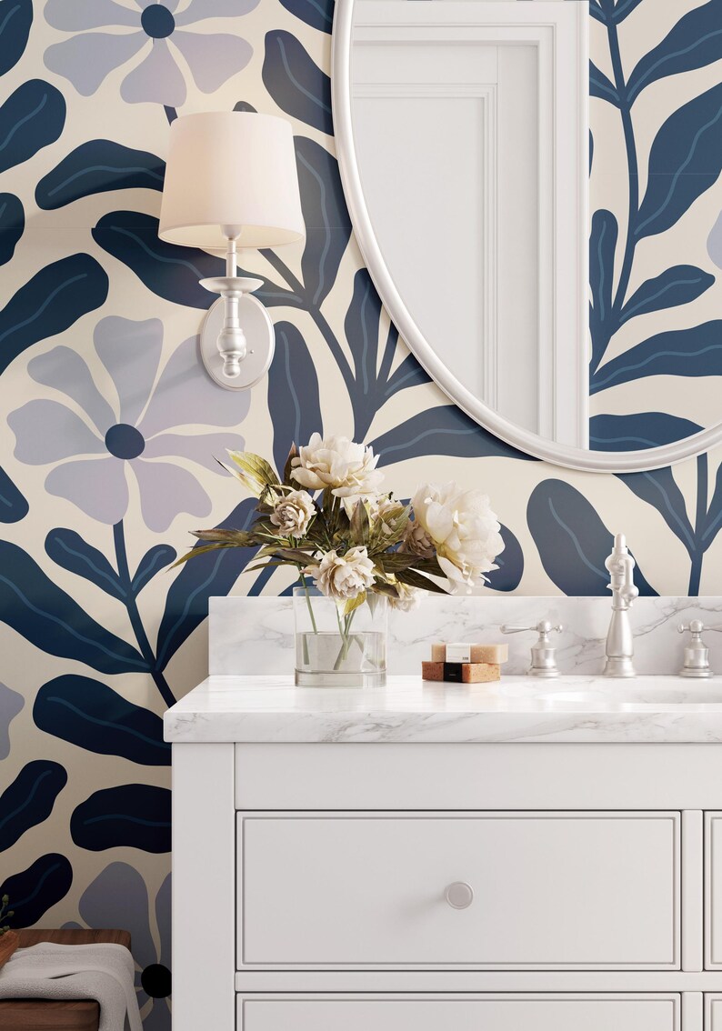 May include: A bathroom scene featuring a white vanity with a marble countertop, a round mirror, and a floral wallpaper in shades of blue and cream. A bouquet of white flowers sits in a clear vase.