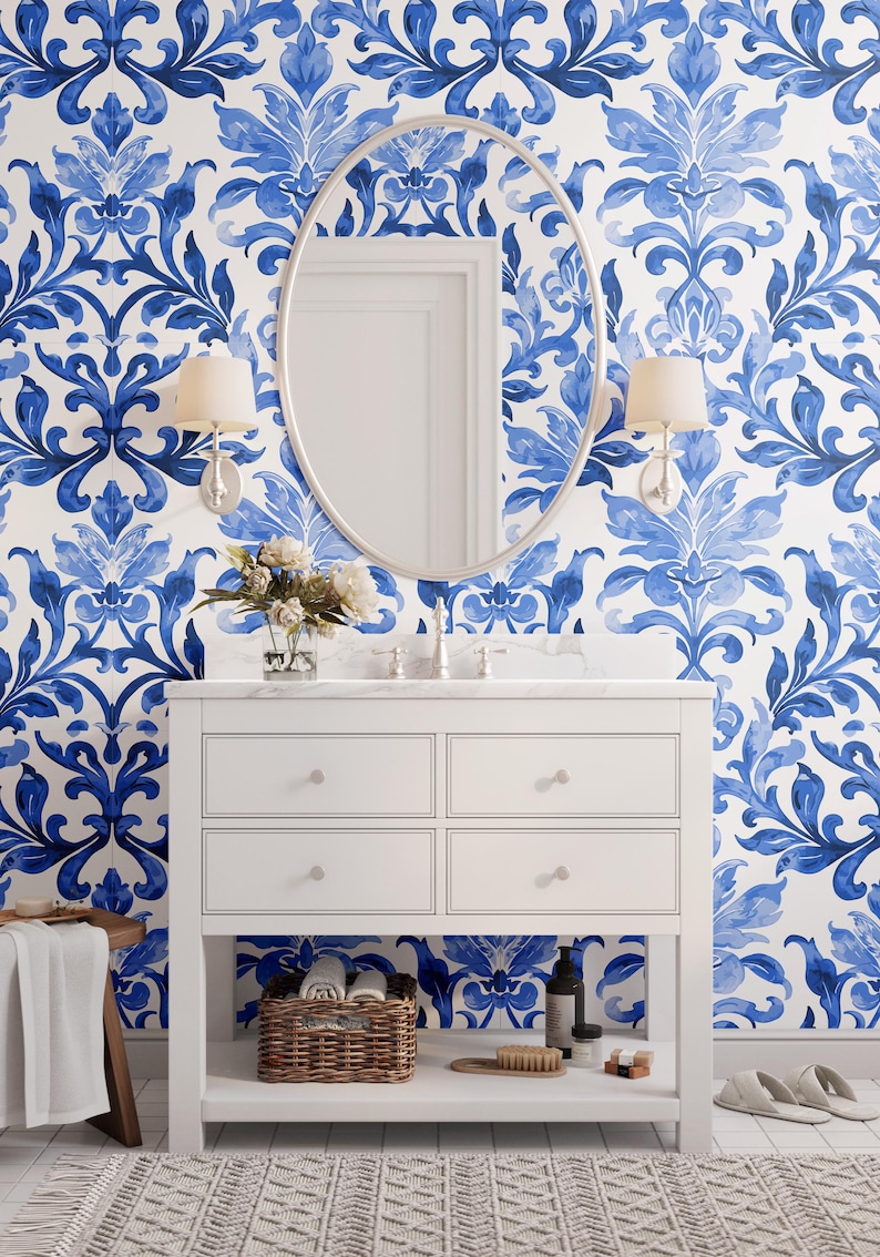 May include: A white bathroom vanity with four drawers and an open bottom shelf, featuring a marble countertop, a silver faucet, and a large oval mirror. The wall is covered in blue and white damask wallpaper with two wall-mounted light fixtures.