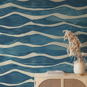 May include: Blue and beige wave pattern wallpaper. The design features a watercolor effect with layered, undulating lines in shades of blue and off-white. The wallpaper creates a serene, coastal aesthetic. A wooden cabinet with a vase of dried plants is in the foreground.