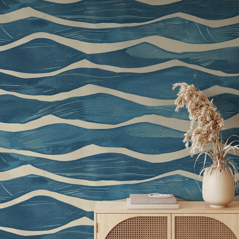 May include: Blue and beige wave pattern wallpaper. The design features a watercolor effect with layered, undulating lines in shades of blue and off-white. The wallpaper creates a serene, coastal aesthetic. A wooden cabinet with a vase of dried plants is in the foreground.