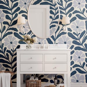 May include: A bathroom scene featuring a white vanity with a marble countertop, an oval mirror, and floral wallpaper in blue and cream. The vanity has four drawers and a lower shelf with a wicker basket and toiletries. Two sconces flank the mirror.