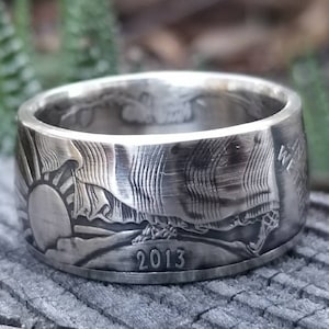 May include: A silver ring made from a 2013 US quarter dollar coin. The ring features an engraved design of a sunrise and a mountain range. The year 2013 is visible on the bottom of the ring.