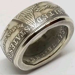 May include: A silver ring crafted from a coin, featuring embossed text and intricate details. The ring's band displays the words "UNITED STATES OF AMERICA" and "IN GOD WE TRUST". The ring has a textured edge and a polished finish.